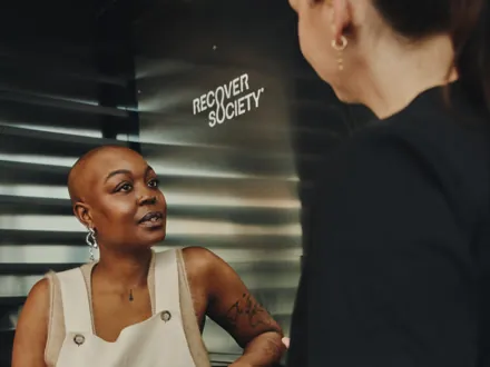 Gast im Gespräch mit einer Mitarbeiterin an der Markenwand von Recover Society